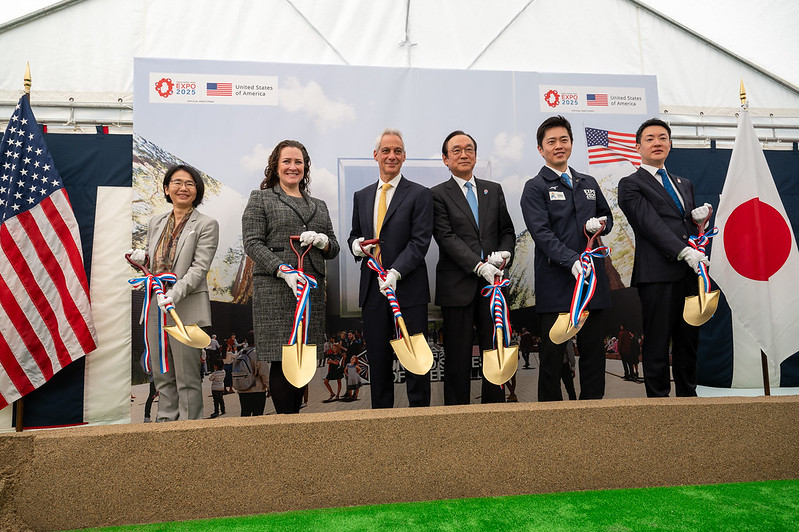 Two women and four men are about to put shovels on the sand whose standing between USA flag on left and Japan flag on right.